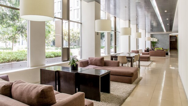 Un moderno vestíbulo de hotel con ventanas amplias, una zona de estar central y un largo pasillo.