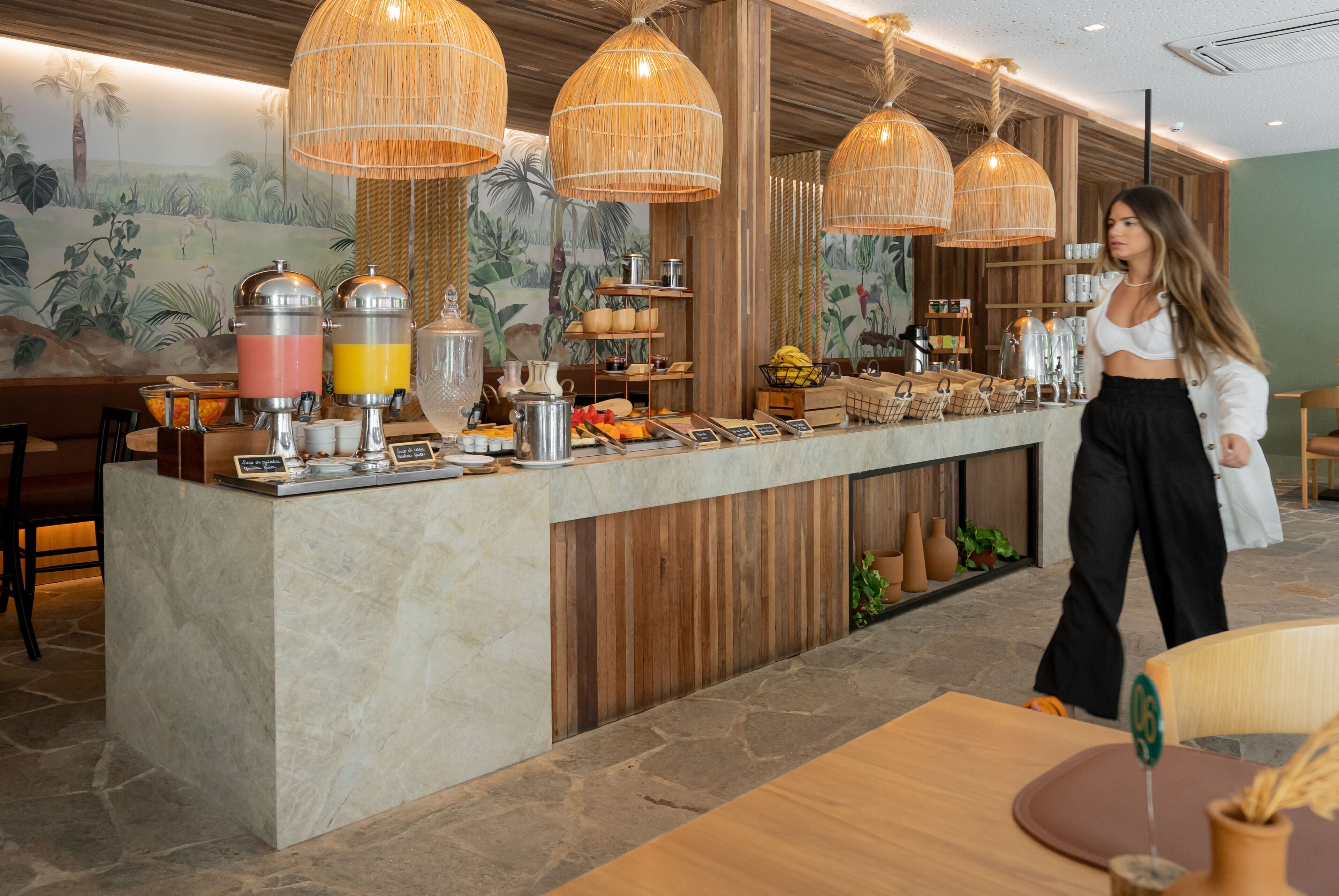 A breakfast bar with a variety of food items and beverages, a mural of a tropical scene, and wicker pendant lights.