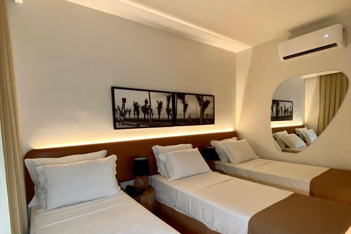 A hotel room with three single beds, a large wall-mounted artwork, a round mirror, and an air conditioning unit.