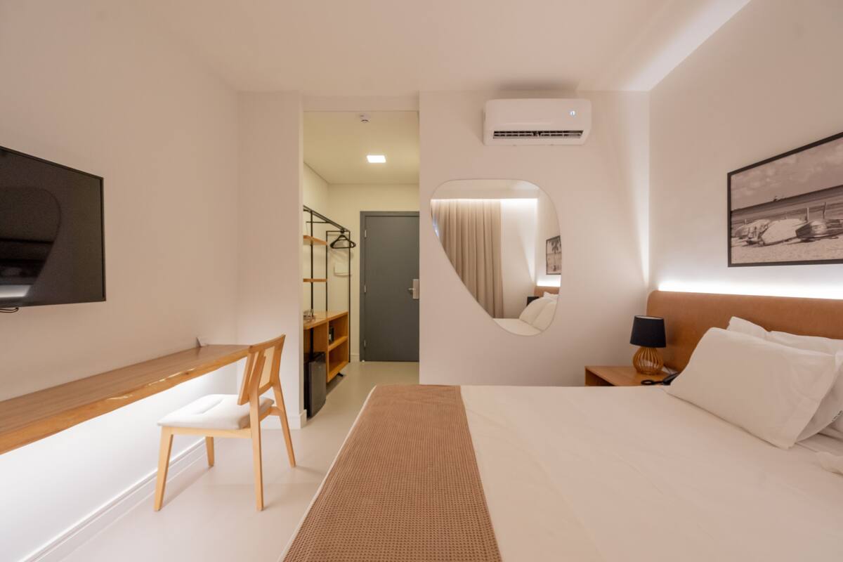A hotel room with a bed, a desk with a chair, a mirror, a TV mounted on the wall, and a framed picture on the wall.