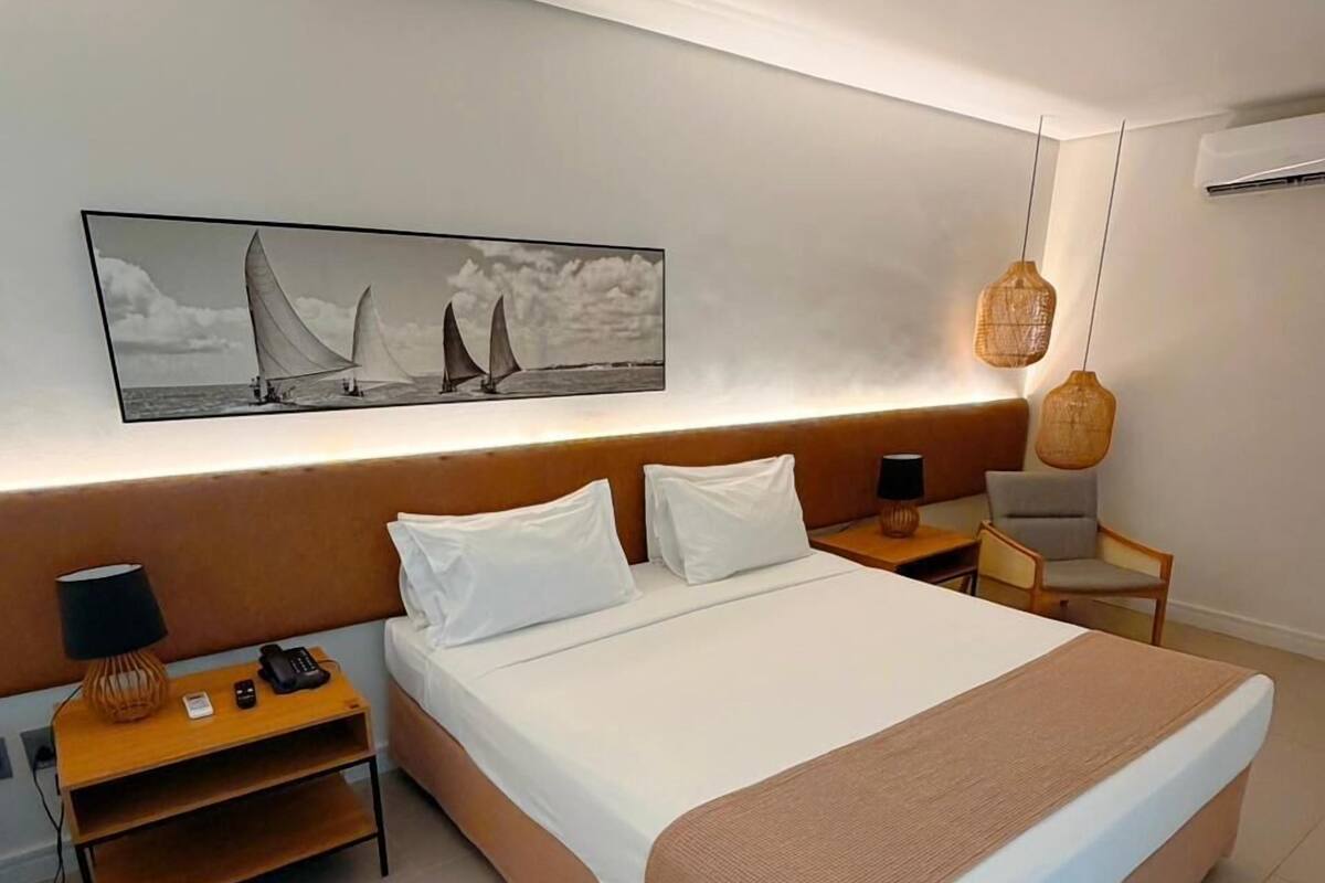 A modern hotel room with a large bed, bedside tables, a lamp, a chair, and a wall-mounted artwork.