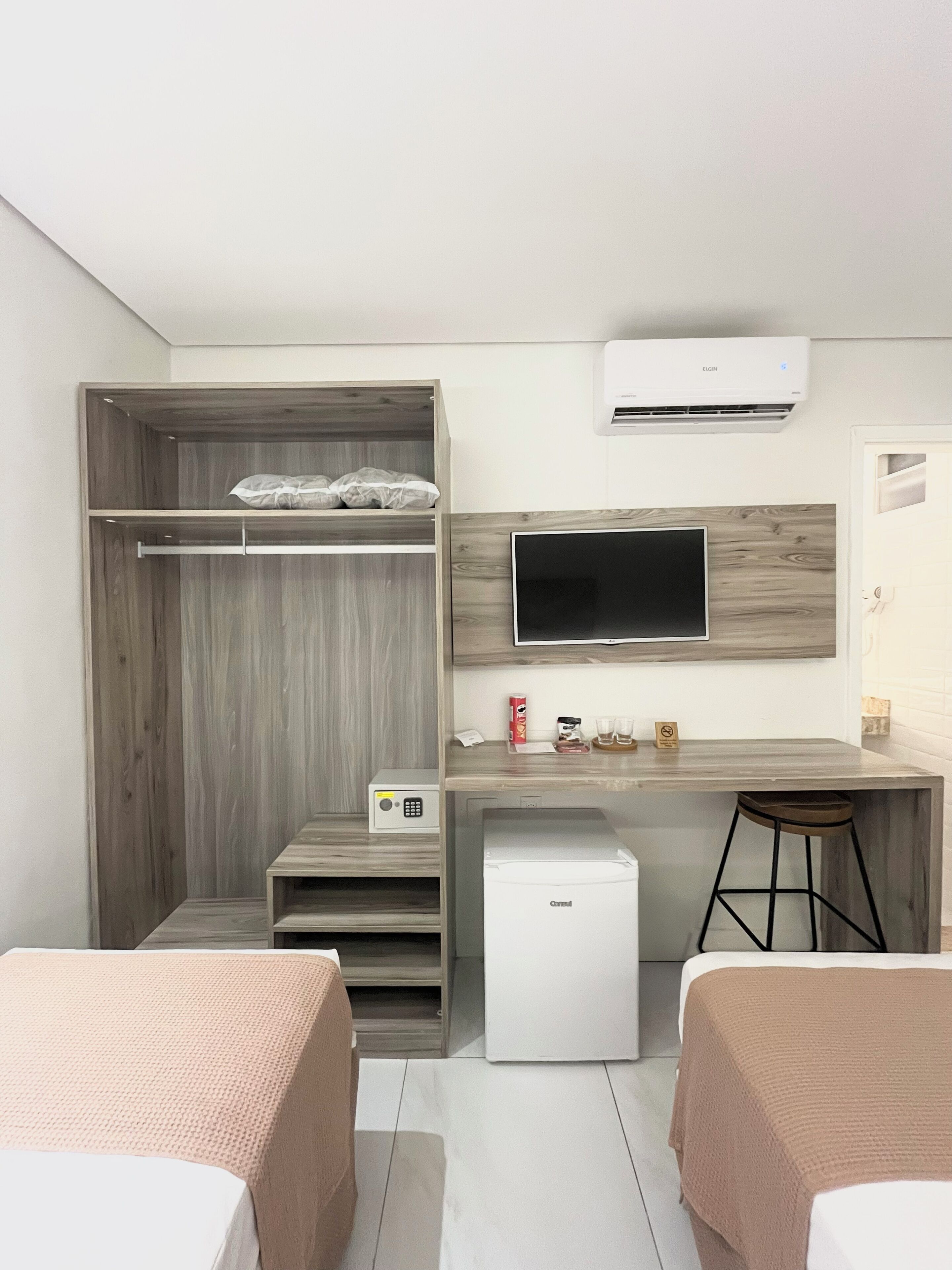 A modern hotel room with a wooden wardrobe, a small kitchenette, a TV mounted on the wall, and a compact refrigerator.