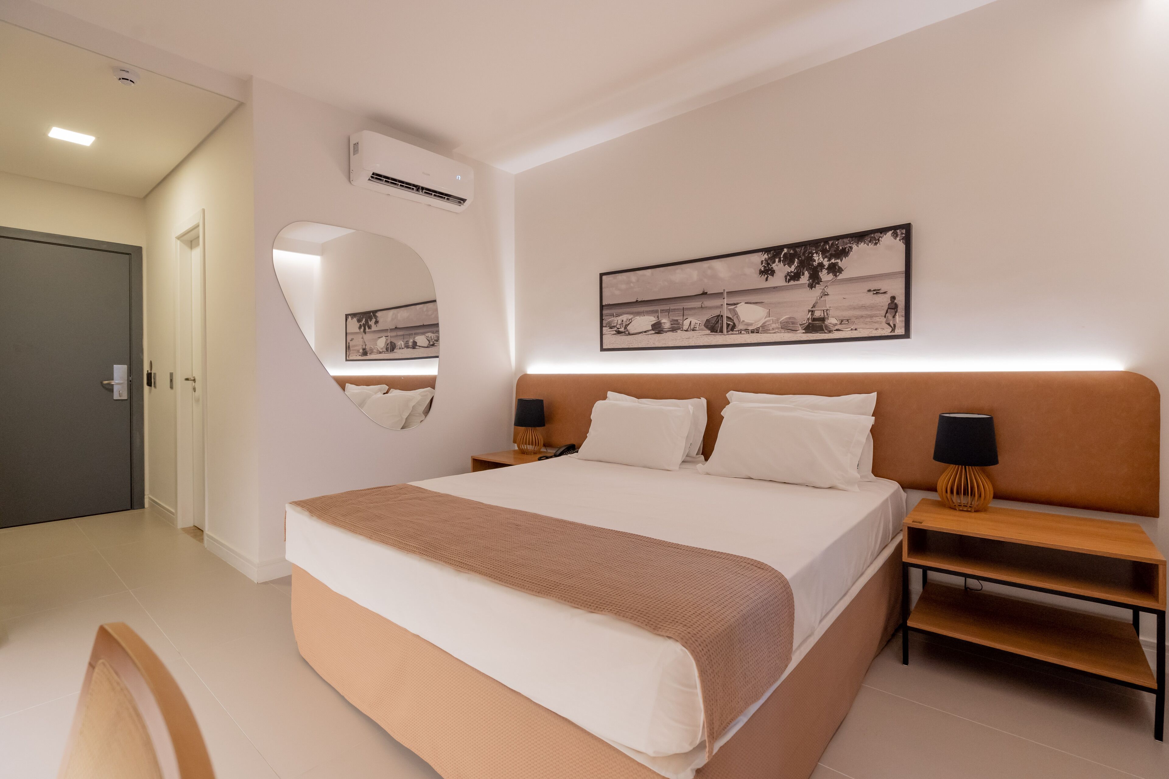Habitación de hotel moderna con una cama grande, una mesita de noche con lámpara, aire acondicionado montado en la pared y un cuadro enmarcado sobre la cama.