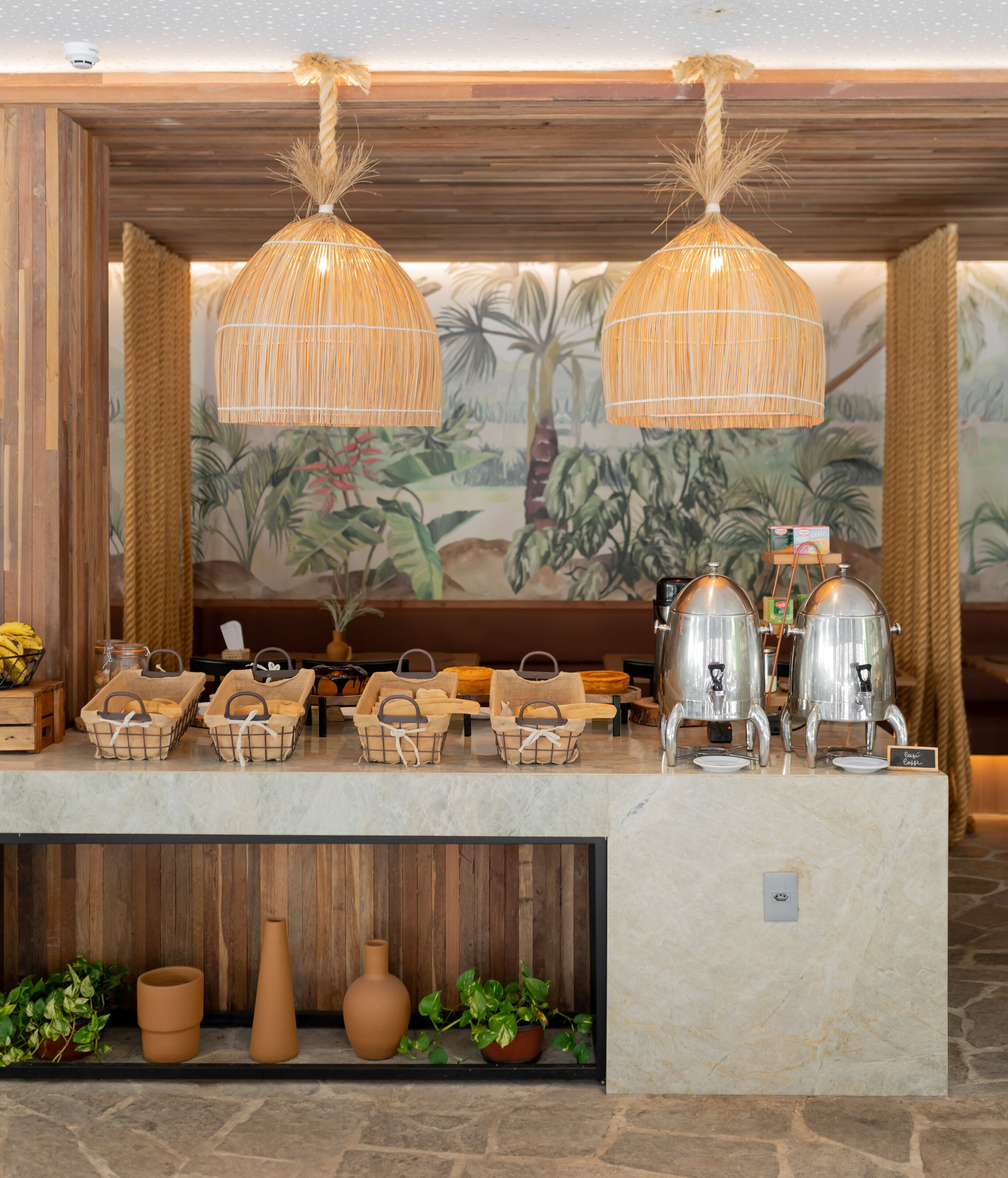 Montaje de buffet con cestas de mimbre, una estación de café y un área de estar con un mural de plantas tropicales.