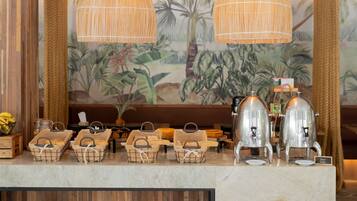 Montaje de buffet con cestas de mimbre, una estación de café y un área de estar con un mural de plantas tropicales.