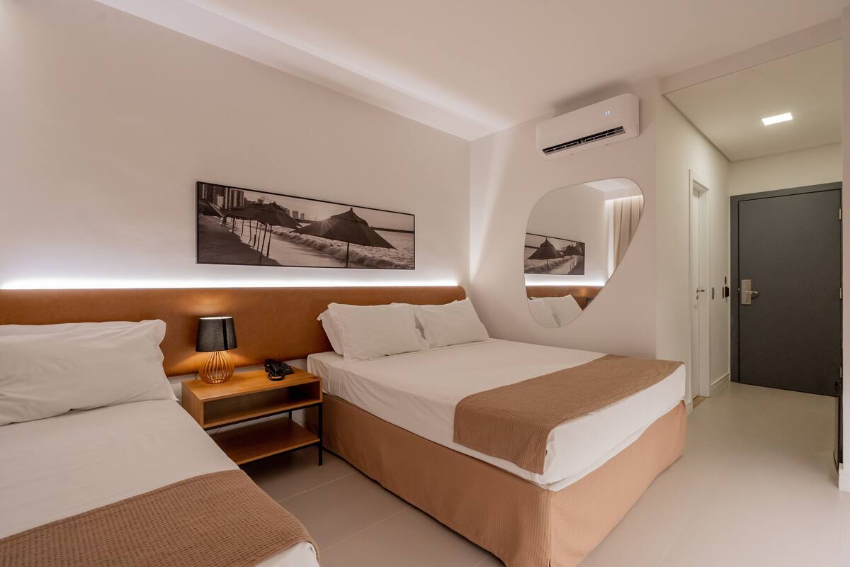 A hotel room with two beds, a bedside table with a lamp, a wall-mounted air conditioner, and a framed picture above the beds.