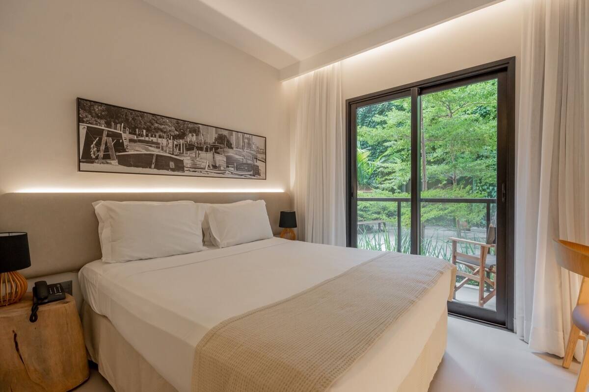 A bedroom with a large bed, a cityscape painting, a wooden nightstand, and a view of greenery through a large window.