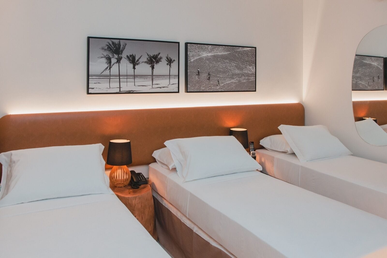 Habitación de hotel con tres camas individuales, una mesita de noche de madera con lámpara y dos cuadros en la pared.
