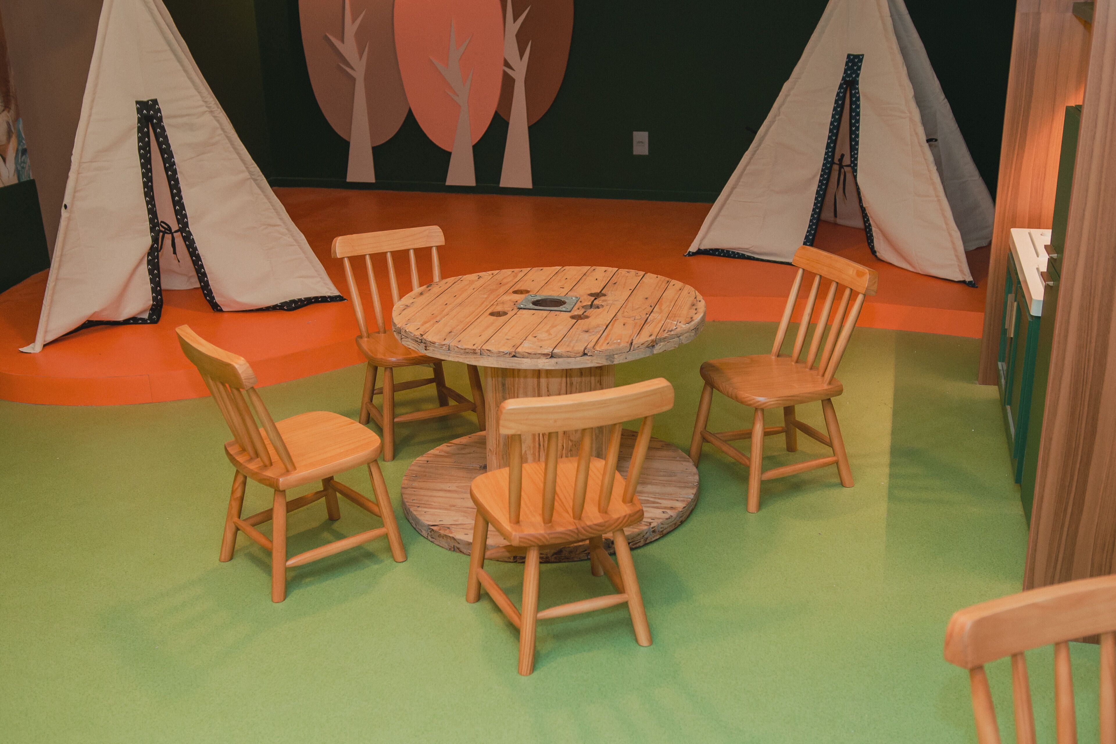 Una habitación con una mesa redonda de madera rodeada por cuatro sillas de madera, dos tiendas blancas al fondo y un suelo cubierto de alfombra verde.