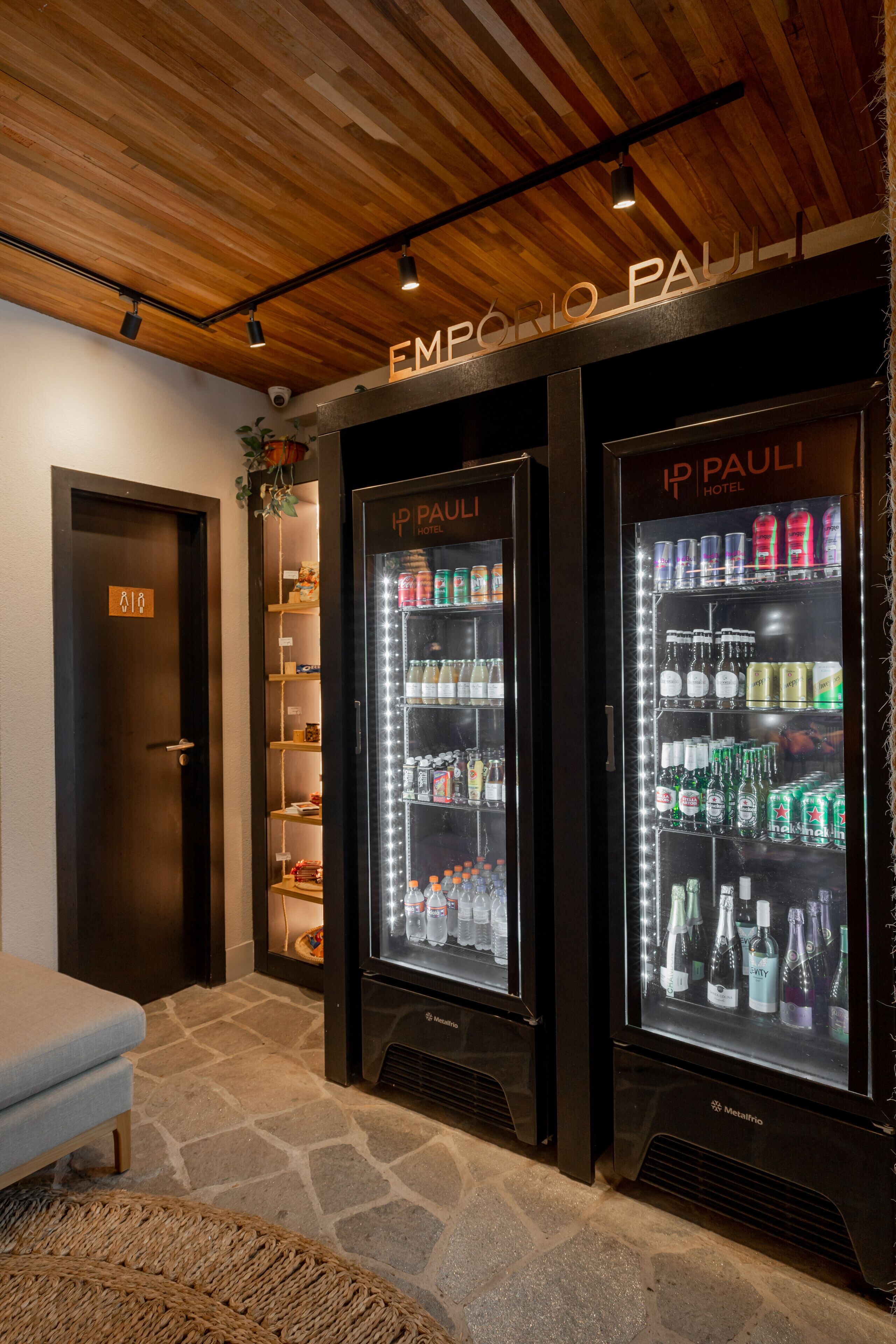 Um saguão de hotel com dois dispensadores de bebidas refrigeradas com a inscrição "IP PAULI HOTEL", piso de ladrilho de pedra, um banco e teto de madeira.
