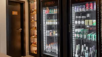Um saguão de hotel com dois dispensadores de bebidas refrigeradas com a inscrição "IP PAULI HOTEL", piso de ladrilho de pedra, um banco e teto de madeira.
