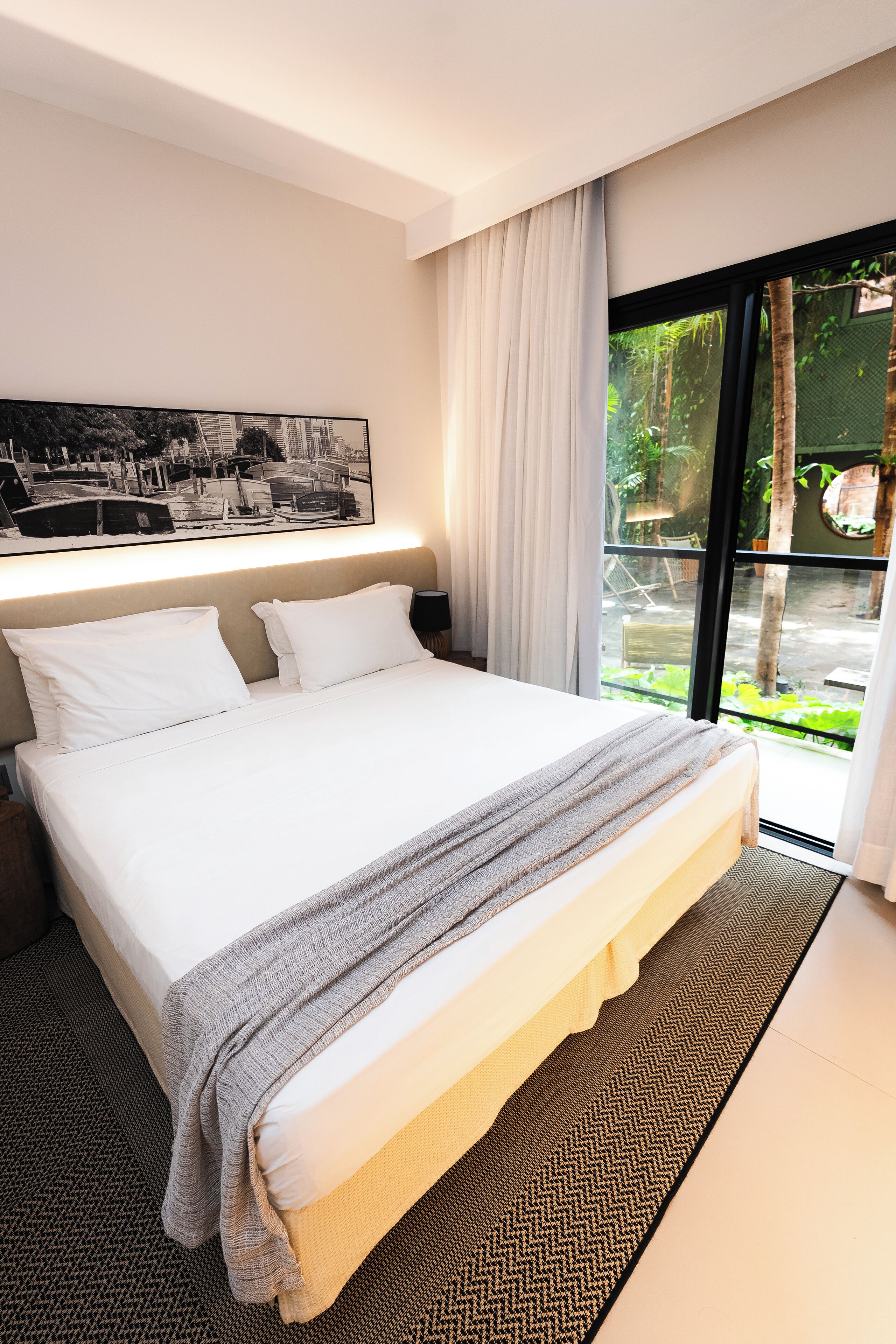 A modern hotel room with a large bed, a headboard with built-in lighting, a view of greenery through a sliding glass door, and a black and white photograph on the wall.
