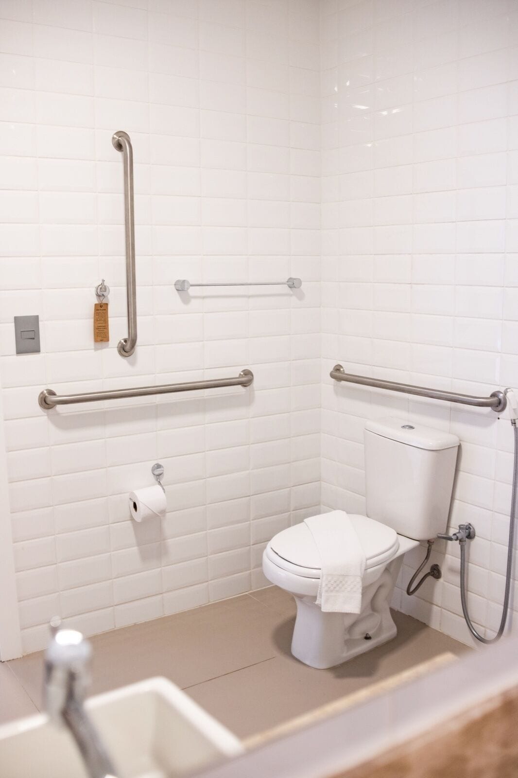 A small, white-tiled bathroom with a toilet, a handrail, and a towel rack.