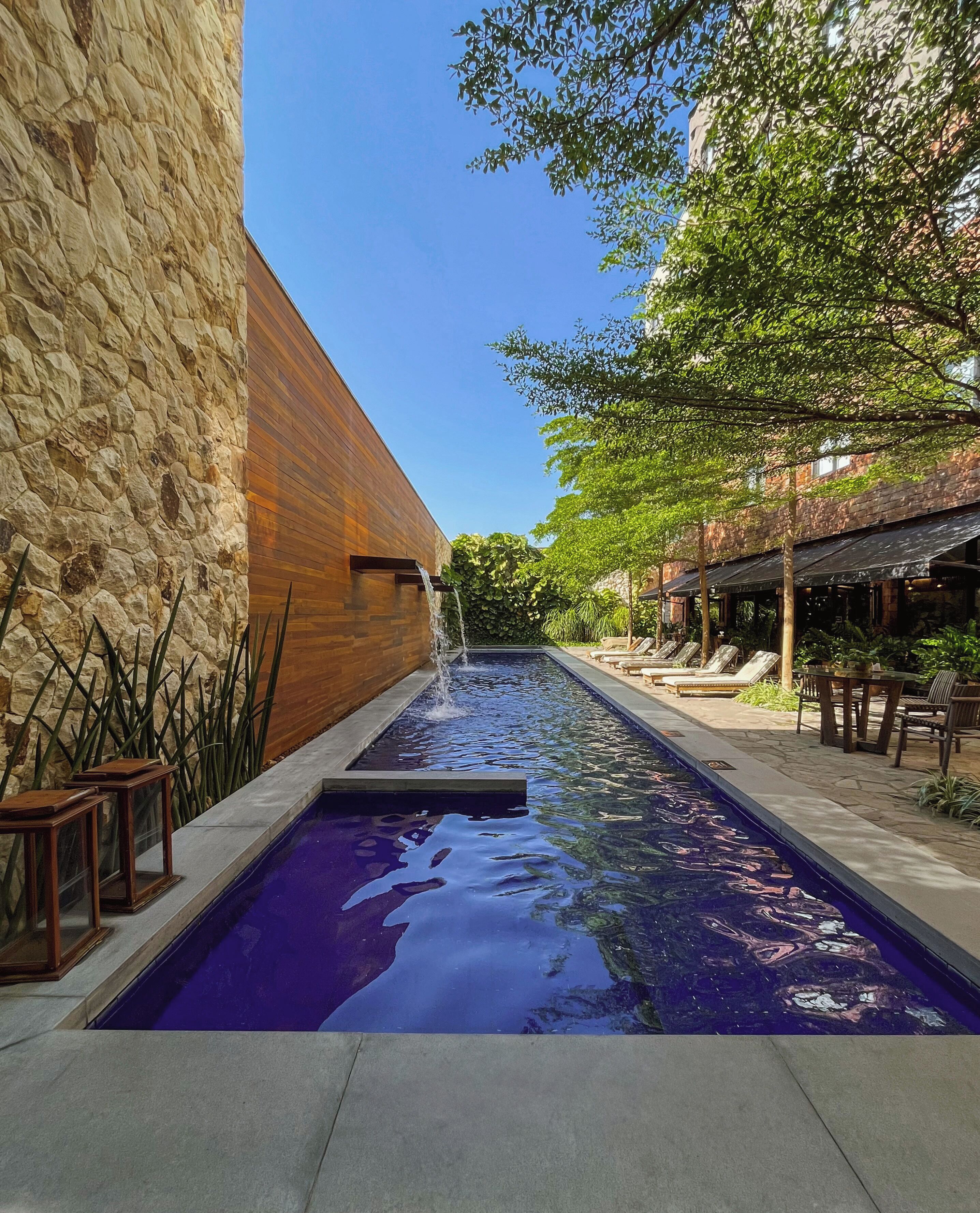 Una piscina con una cascada, rodeada de muros de piedra y paneles de madera, con mobiliario de exterior y abundante vegetación.