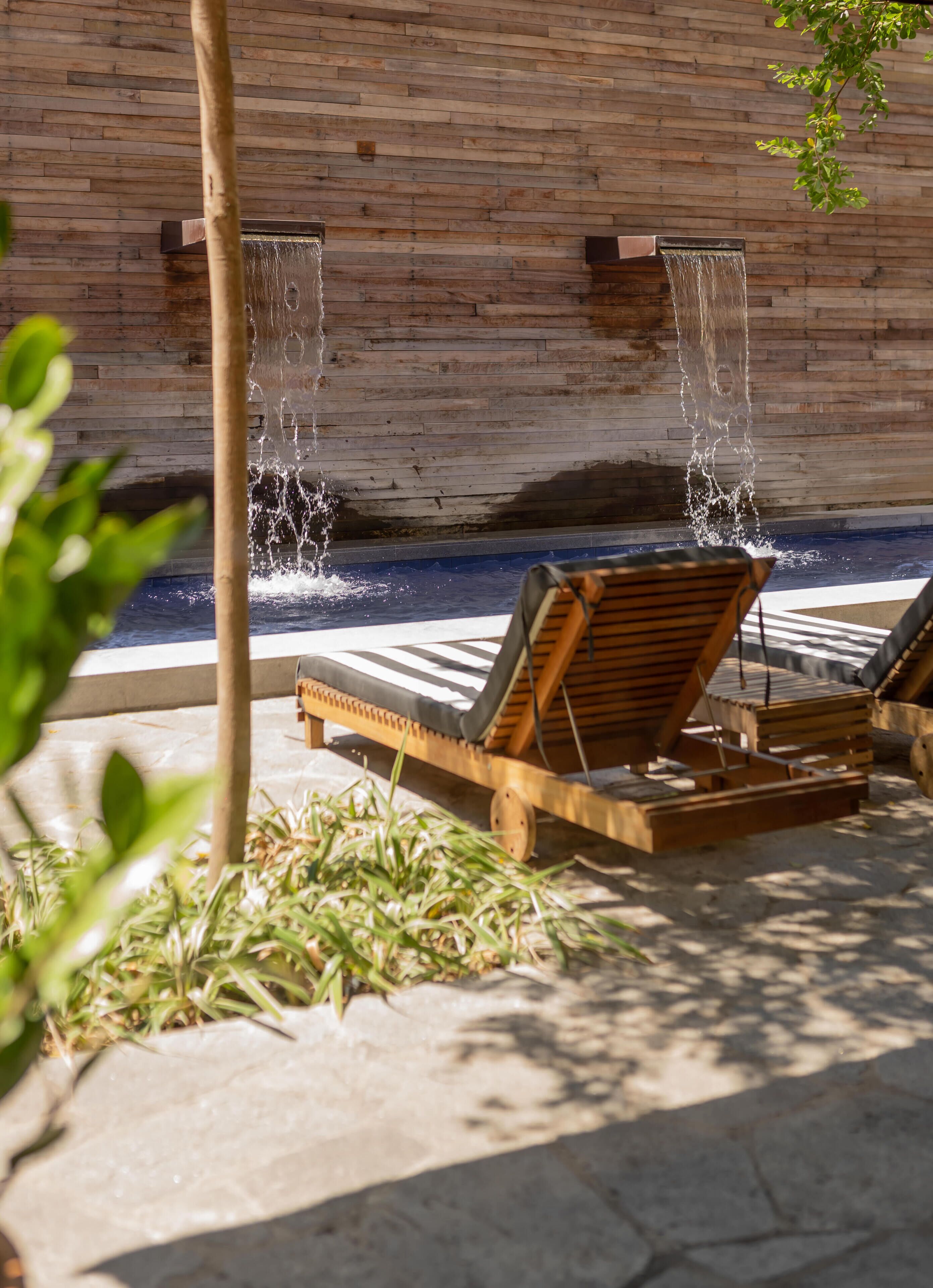 A pool area with two wooden lounge chairs, a wooden wall, and a water feature.
