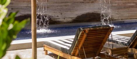 A pool area with two wooden lounge chairs, a wooden wall, and a water feature.