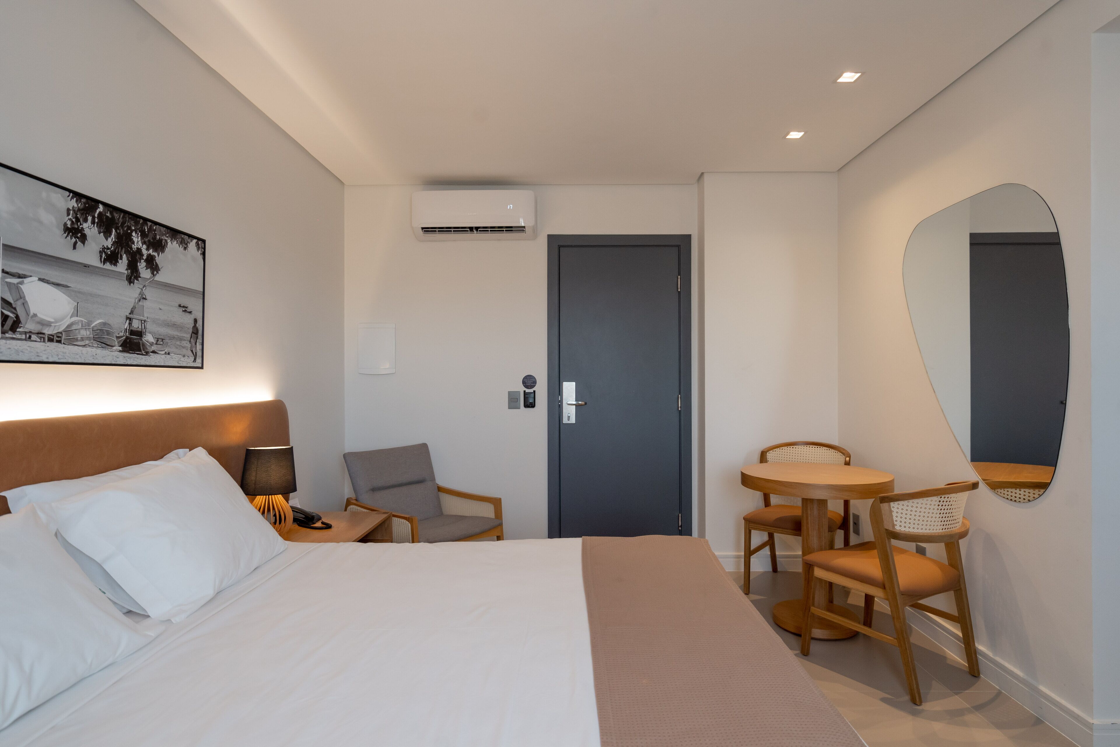 Habitación de hotel con cama, escritorio con silla, mesa redonda con sillas, espejo montado en la pared y una puerta.