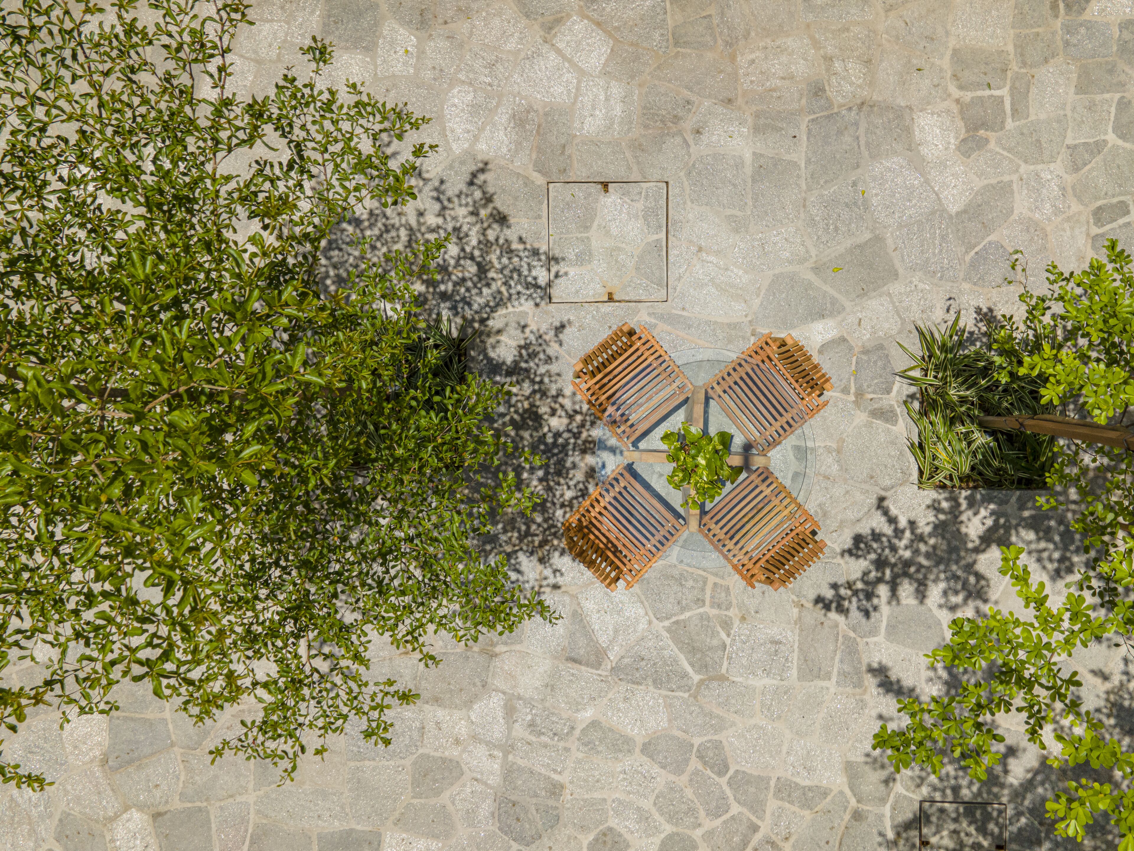 Une terrasse en pierre avec une table ronde et quatre chaises en bois, entourée de verdure.