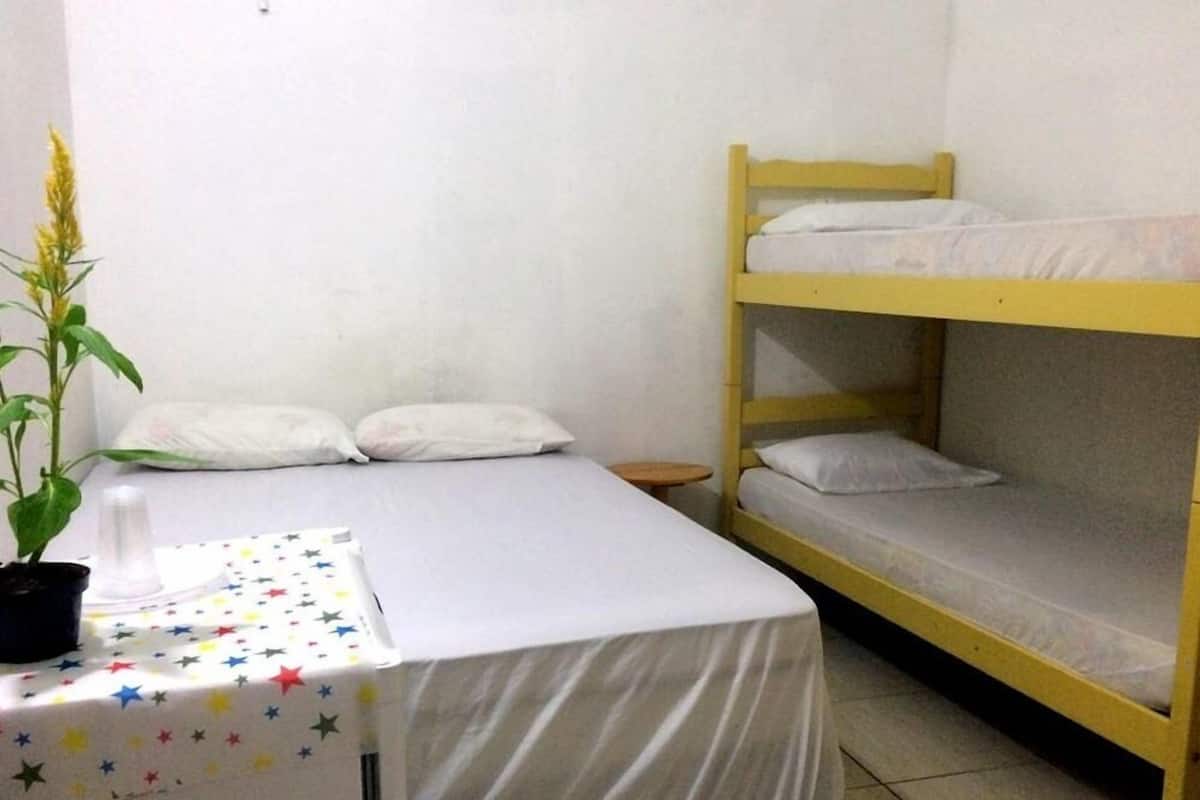 Une petite pièce simple, avec un lit superposé, un lit simple, une petite table recouverte d’une nappe à motifs étoilés et une plante en pot.