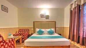 Habitación de hotel con una cama de madera, un sillón rojo estampado, una mesa de centro de madera y una mesita con una botella de agua.