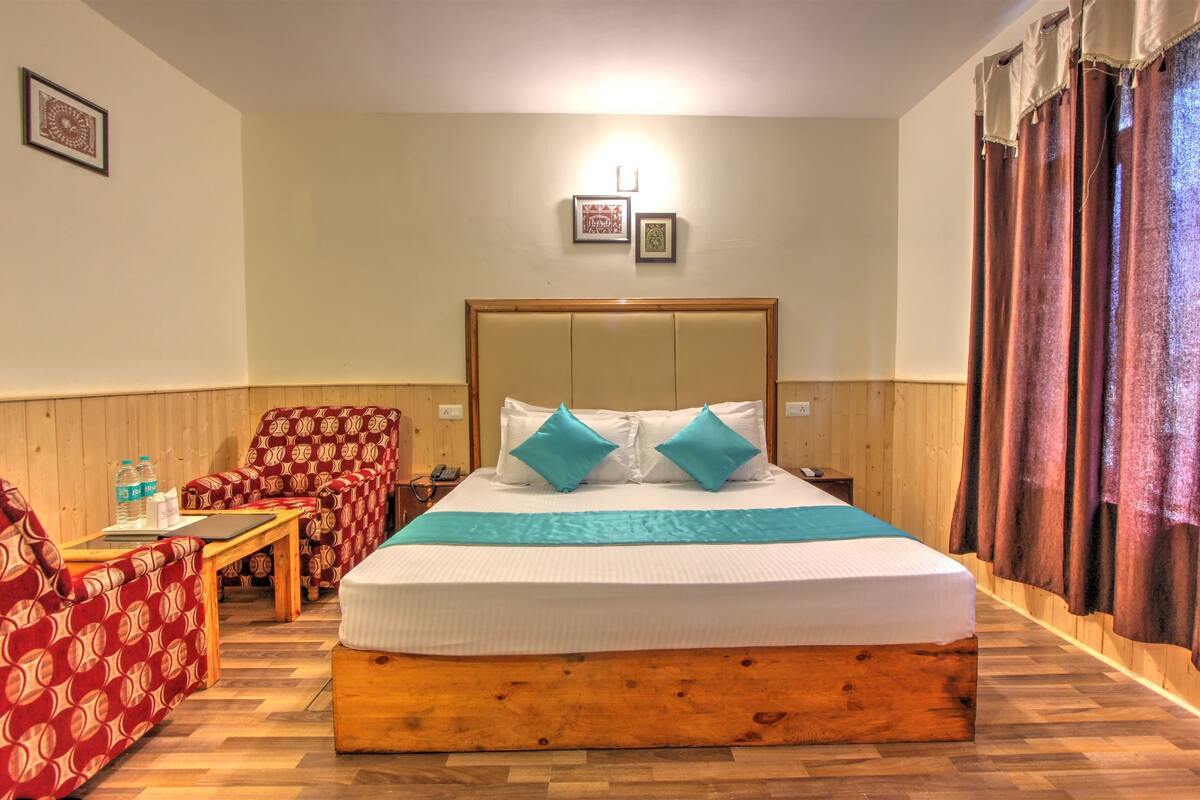 A hotel room with a wooden bed, a red patterned armchair, a wooden coffee table, and a small table with a bottle of water.