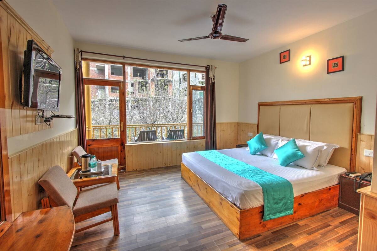 A bedroom with a large bed, wooden headboard, a ceiling fan, a TV mounted on the wall, and a window with a view of buildings.