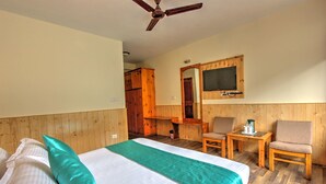 Habitación de hotel con cama, dos sillas, una mesita, ventilador de techo y televisor montado en la pared.