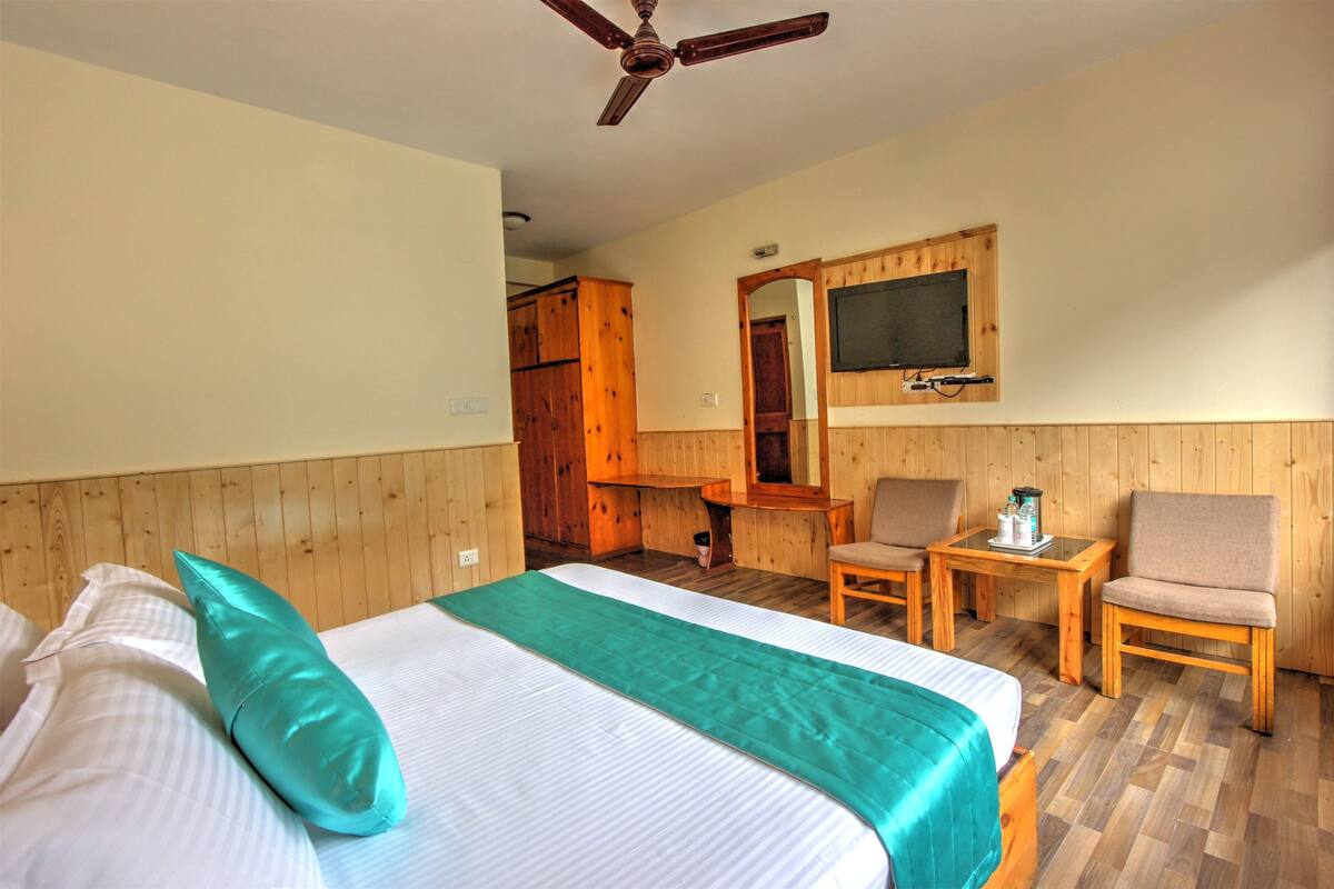 A hotel room with a bed, two chairs, a small table, a ceiling fan, and a television mounted on the wall.