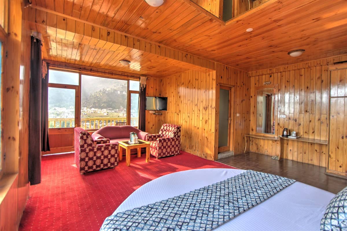 A wooden room with a bed, a TV, a balcony with a view, and two patterned chairs.
