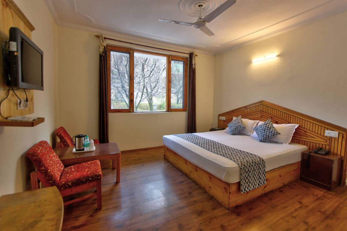 A bedroom with a wooden bed, a desk, a chair, a television, and a window with curtains.