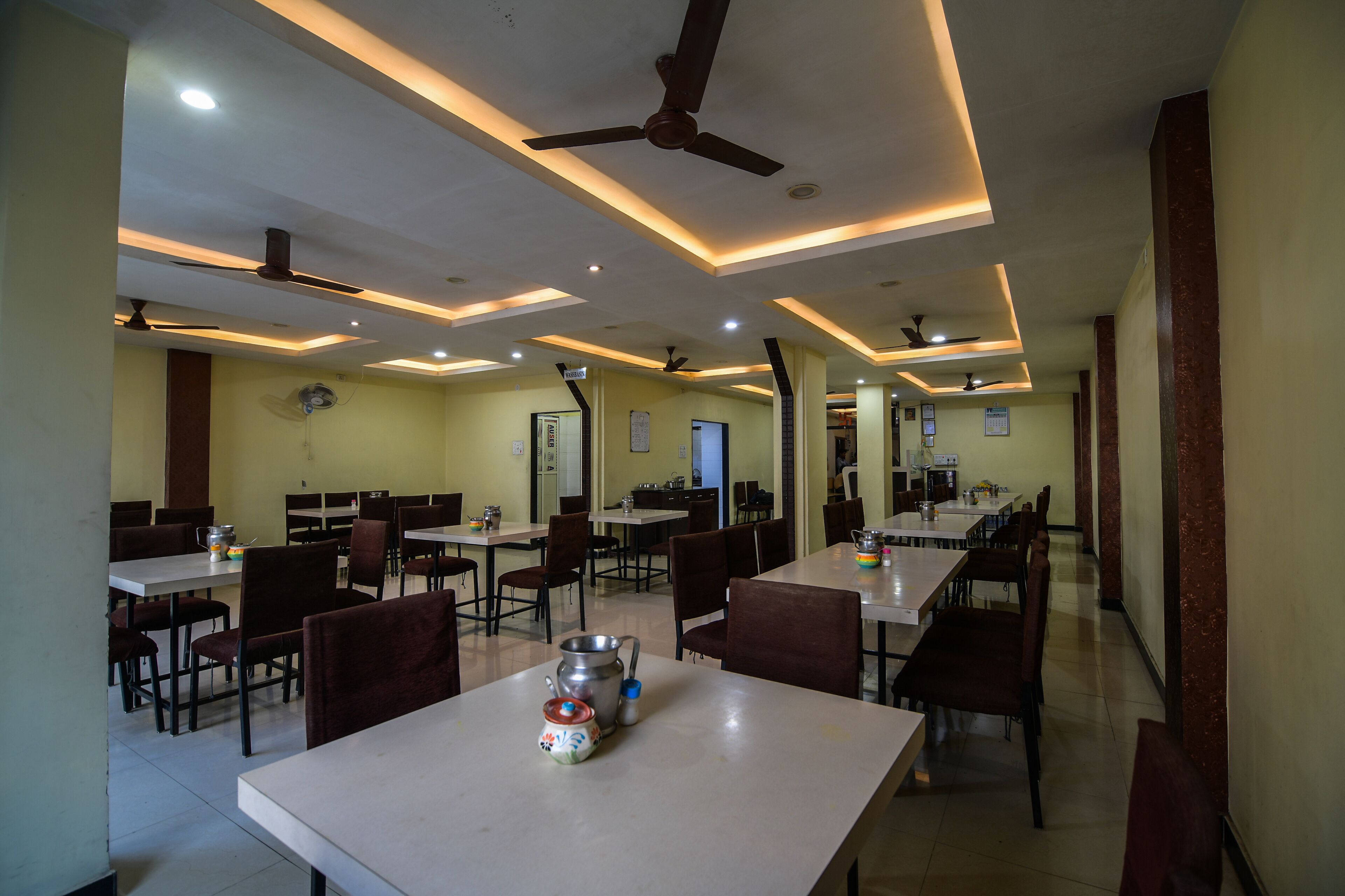 A dining area with multiple tables and chairs, ceiling fans, recessed lighting, and a kitchen area in the background.