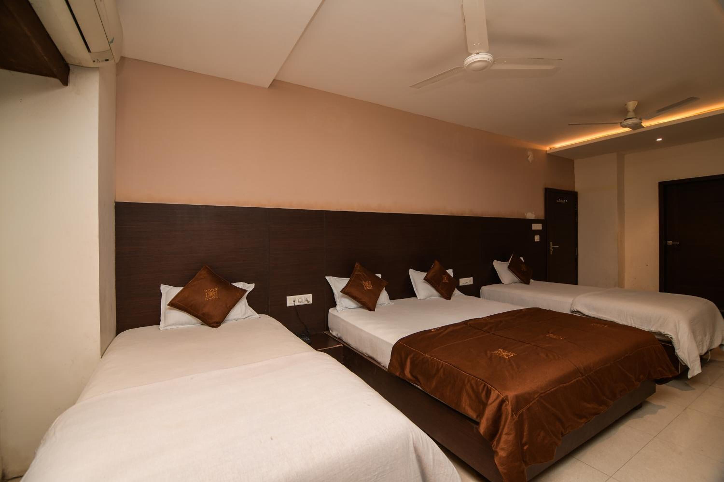 Habitación de hotel con tres camas, cabecera oscura, ventilador de techo y una puerta con manija.