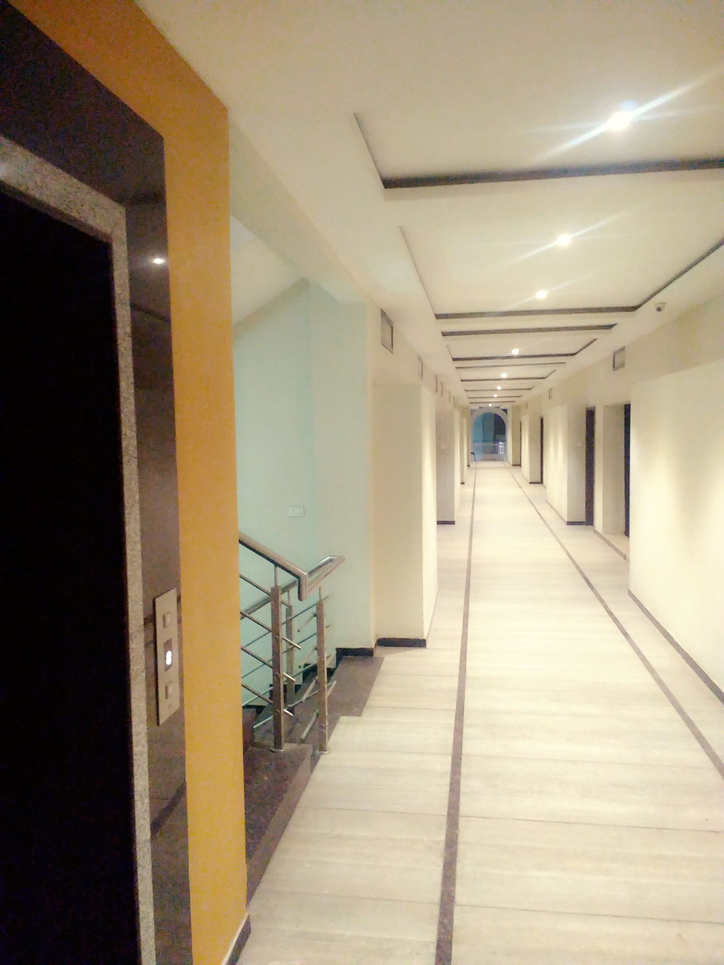 Un long couloir étroit, avec un escalier sur la gauche, menant à un palier. Le couloir est bordé d’une série de portes sur la droite, et le plafond est doté d’un éclairage encastré.