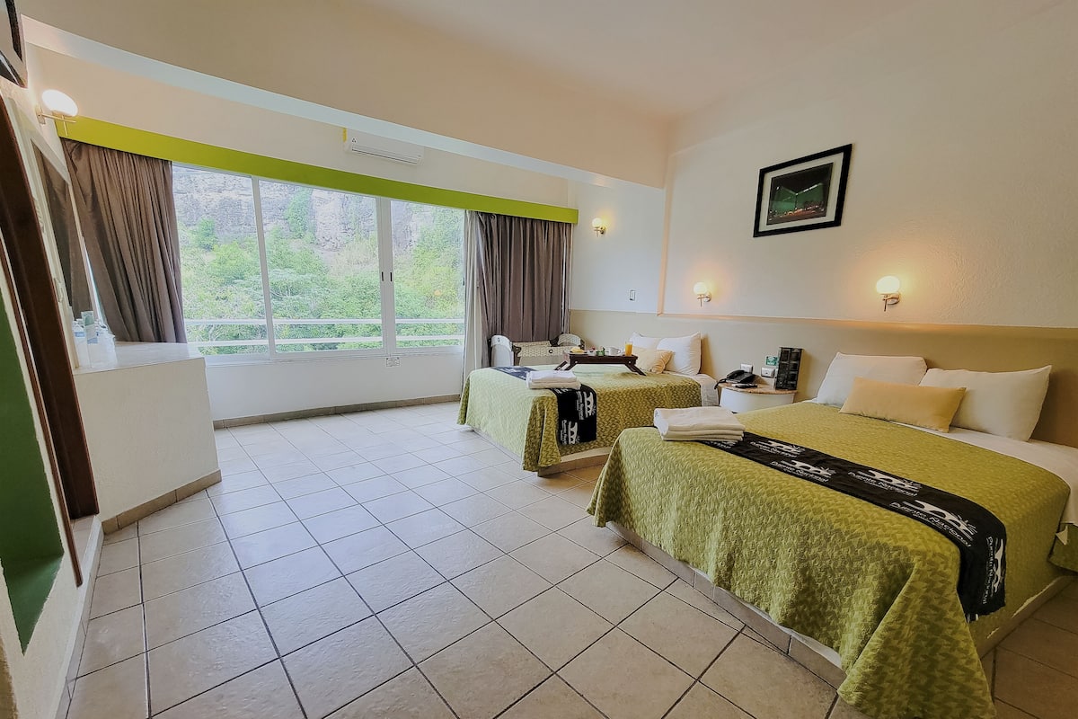 A hotel room with two beds, a balcony, a window with curtains, a bedside table with a lamp, and a wall-mounted picture.