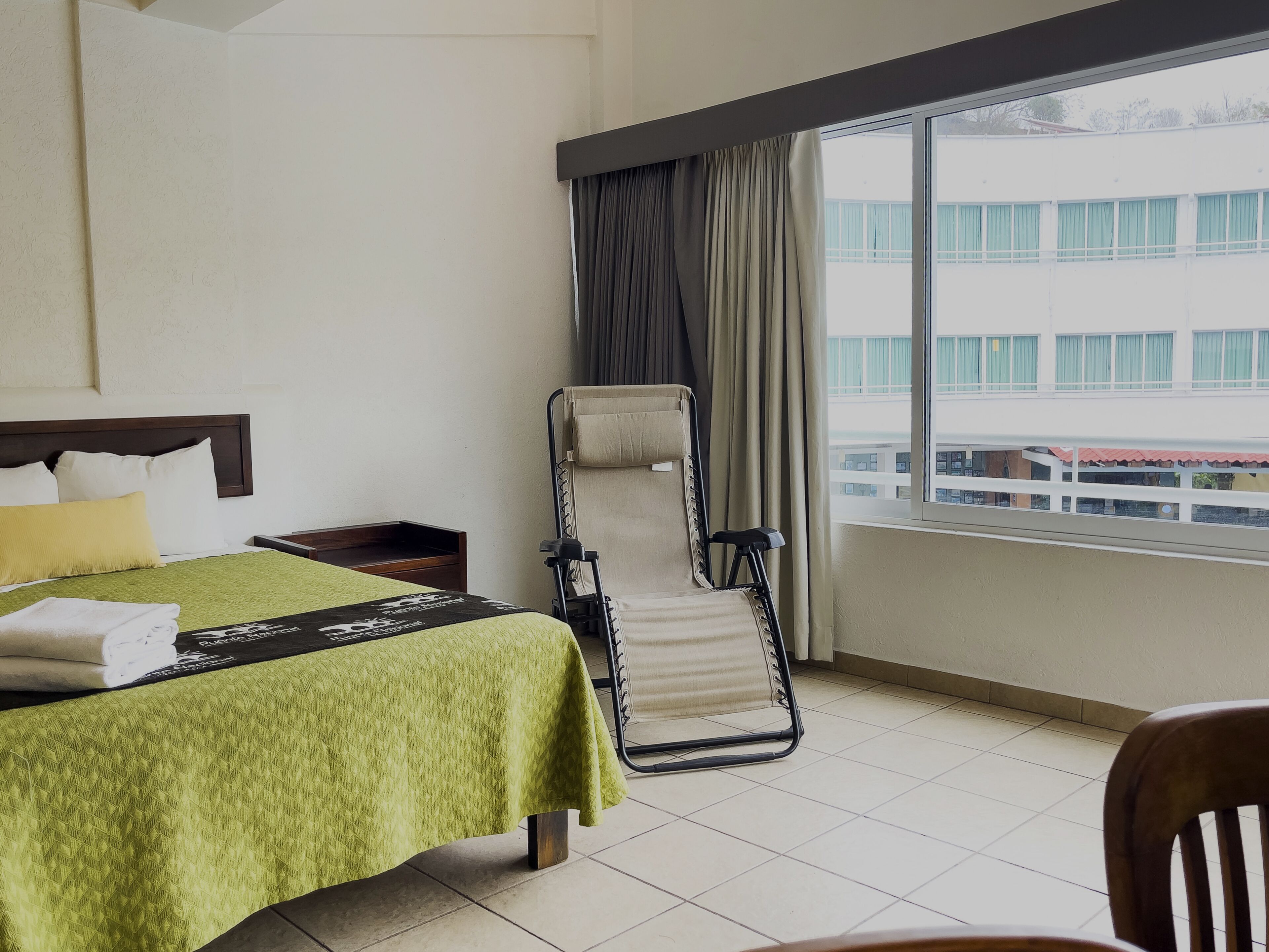 A hotel room with a bed, a chair, a window with curtains, and a view of buildings outside.