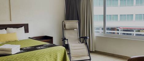 A hotel room with a bed, a chair, a window with curtains, and a view of buildings outside.
