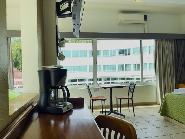 Una habitación de hotel con un ventanal grande, una cafetera sobre la mesa, dos sillas, una cama con cubrecama verde y un equipo de aire acondicionado en la pared.