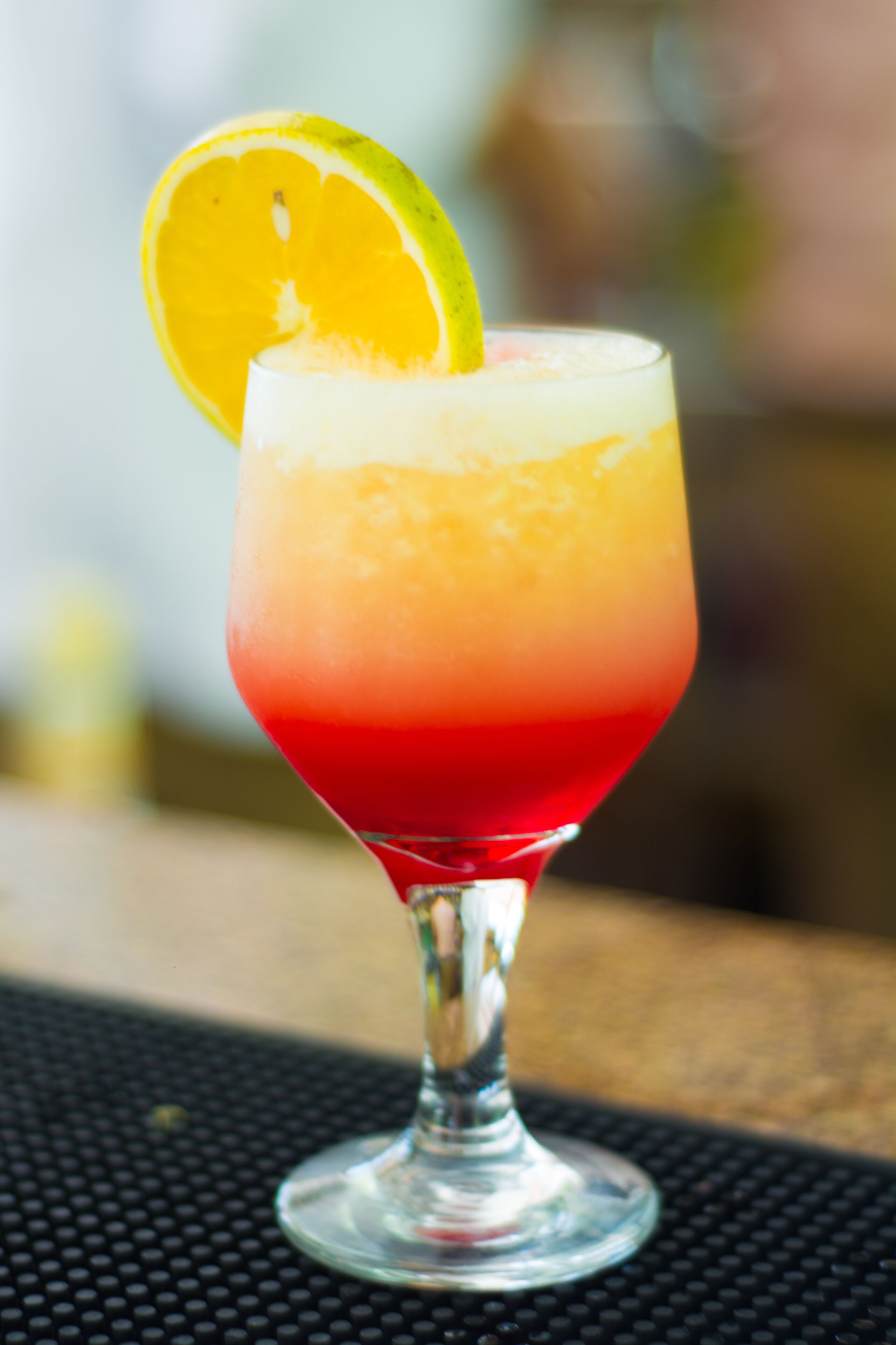 Un cocktail servi dans un verre à pied, avec un dégradé de couleurs allant du rouge au jaune, et décoré d’une tranche d’orange.