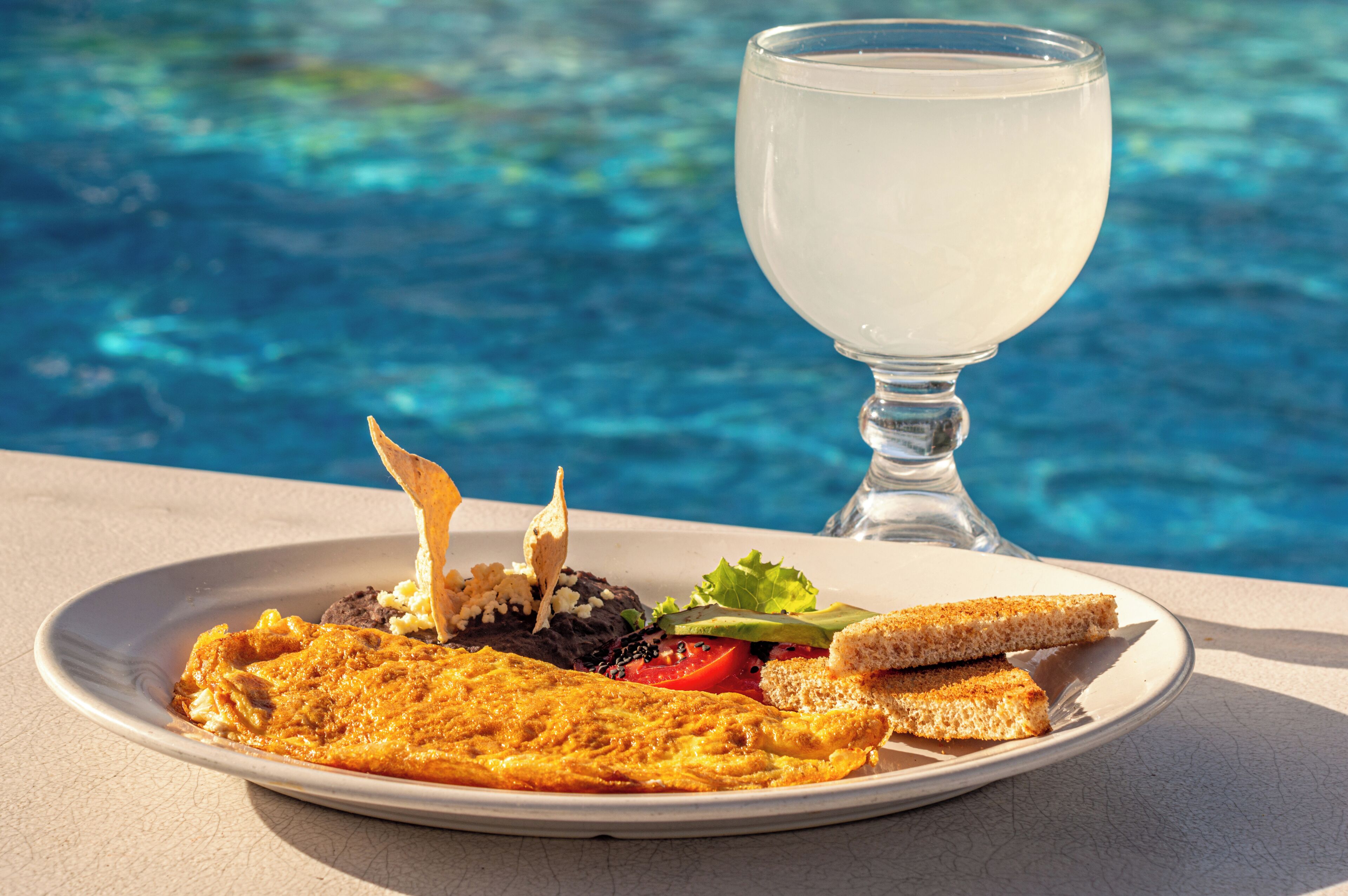 A plate of food with toast, salad, and a glass of white beverage by a pool.