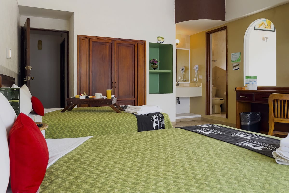 A hotel room with two beds, a wooden wardrobe, a desk with a chair, a bathroom with a mirror, and a small shelf with a vase.