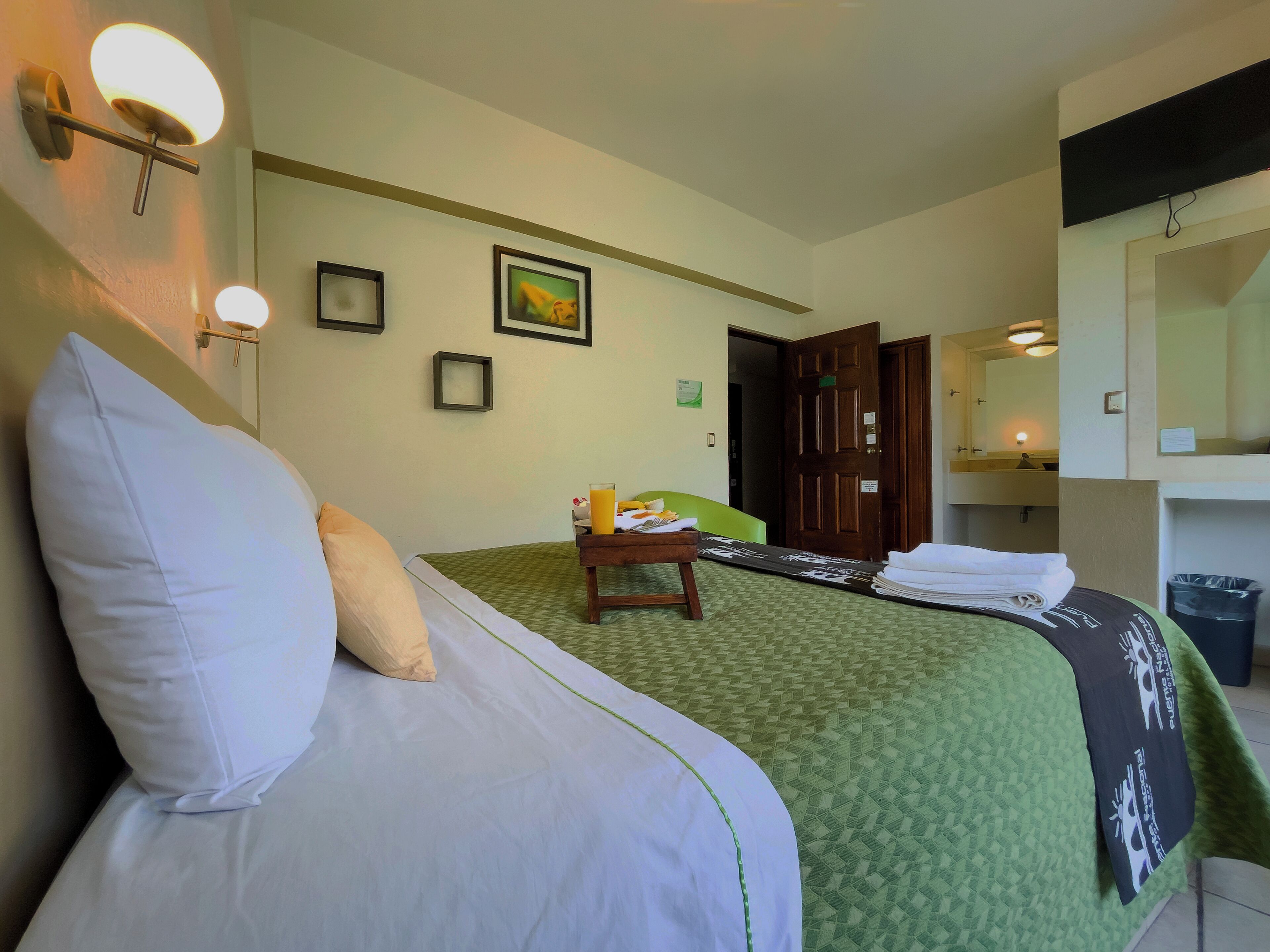Una habitación de hotel con una cama, una mesita de noche con un vaso de jugo de naranja, una colcha de cama estampada en verde, un cuadro en la pared y una puerta que da a otra habitación.