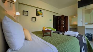 Una habitación de hotel con una cama, una mesita de noche con un vaso de jugo de naranja, una colcha de cama estampada en verde, un cuadro en la pared y una puerta que da a otra habitación.