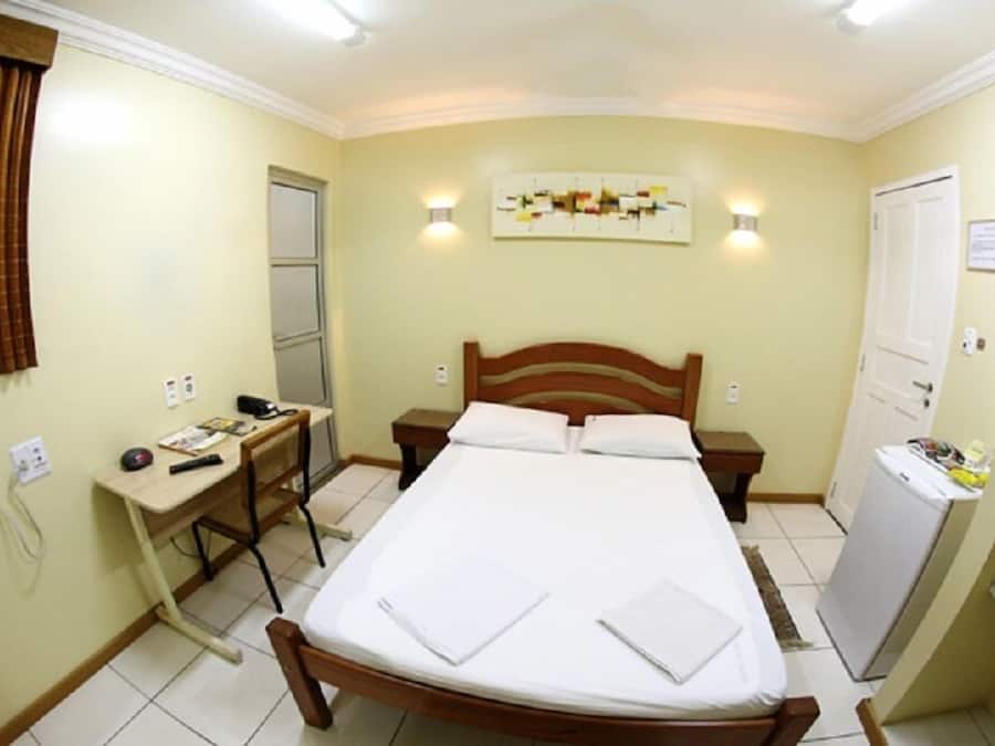 A small, well-lit bedroom with a single bed, a desk, and a refrigerator.