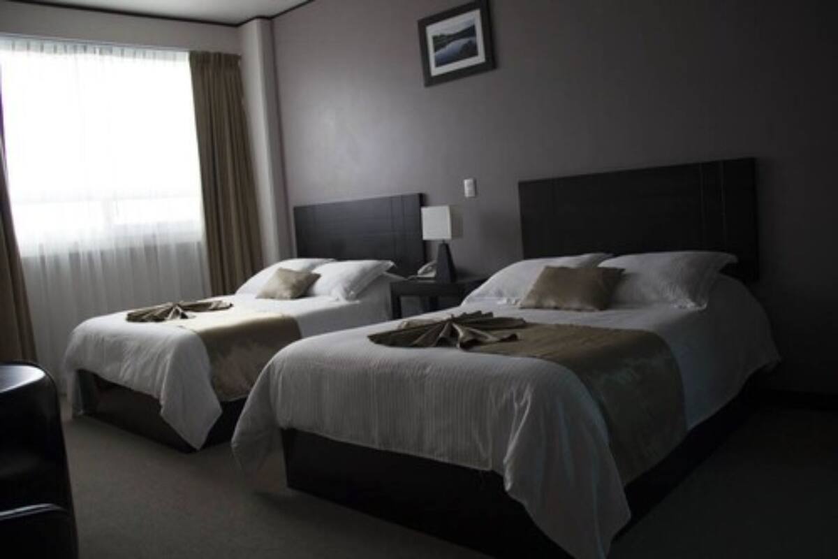 Habitación de hotel con dos camas, una mesita de noche, una lámpara y una ventana con cortinas.
