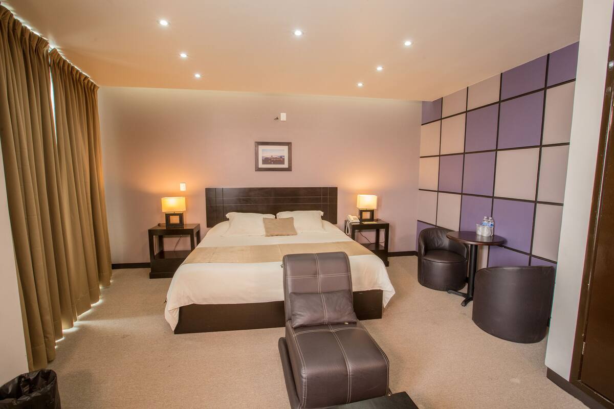 Habitación de hotel con una cama grande, dos lámparas de noche, un cabecero, una mesita, una silla y un cubo de basura.