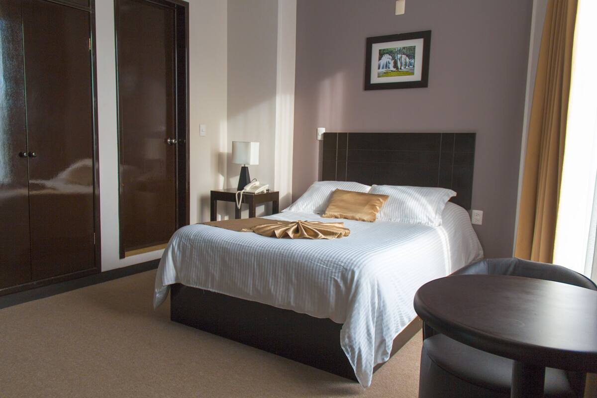 Habitación de hotel con una cama, una mesita de noche con lámpara, un teléfono y un cuadro en la pared.