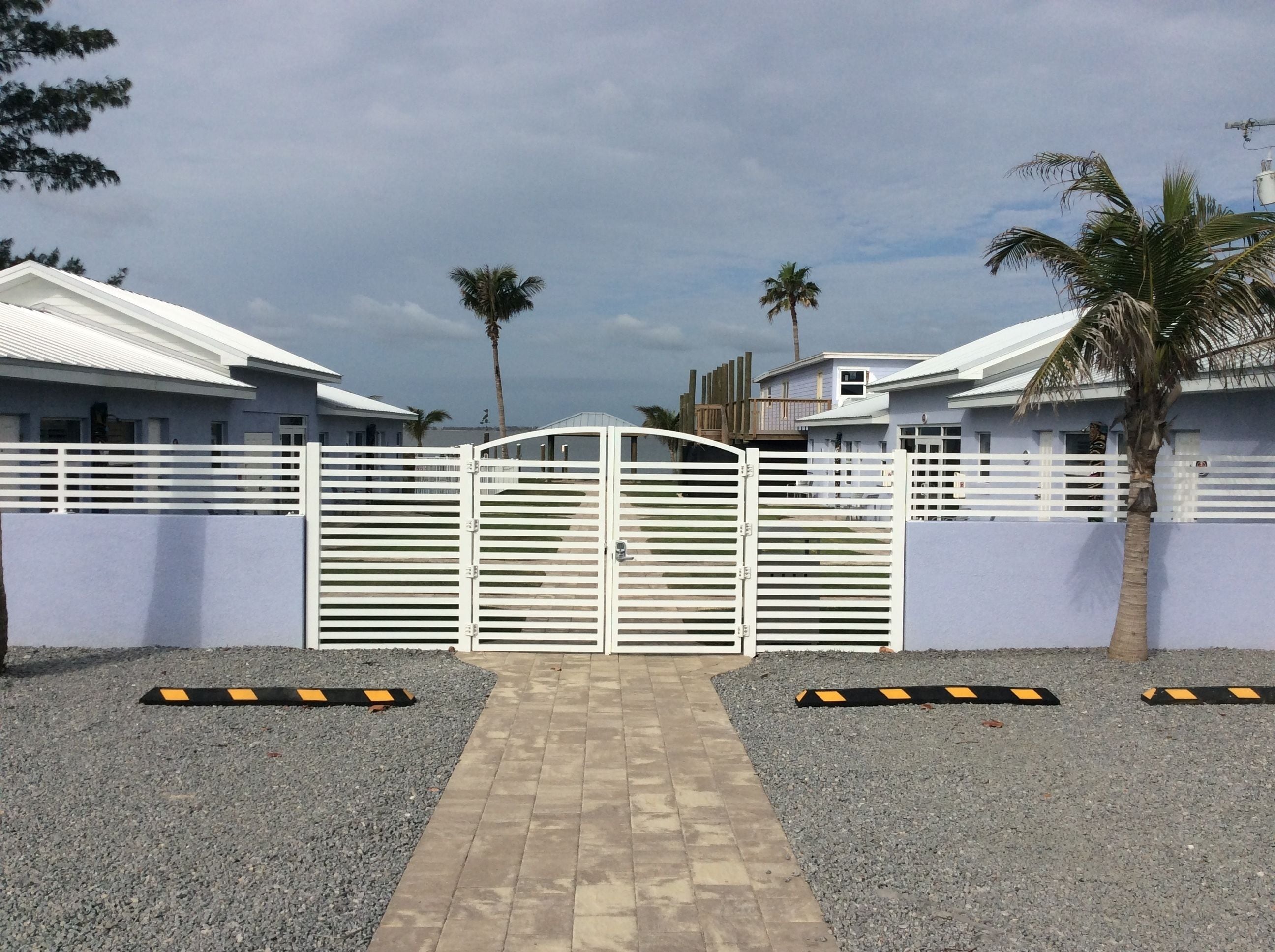 Une entrée fermée par un portail, avec un chemin pavé menant à un bâtiment doté d’un toit blanc et d’un mur gris.