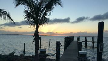 Un muelle de madera que se adentra en aguas tranquilas, con una palmera a la izquierda y un atardecer de fondo.