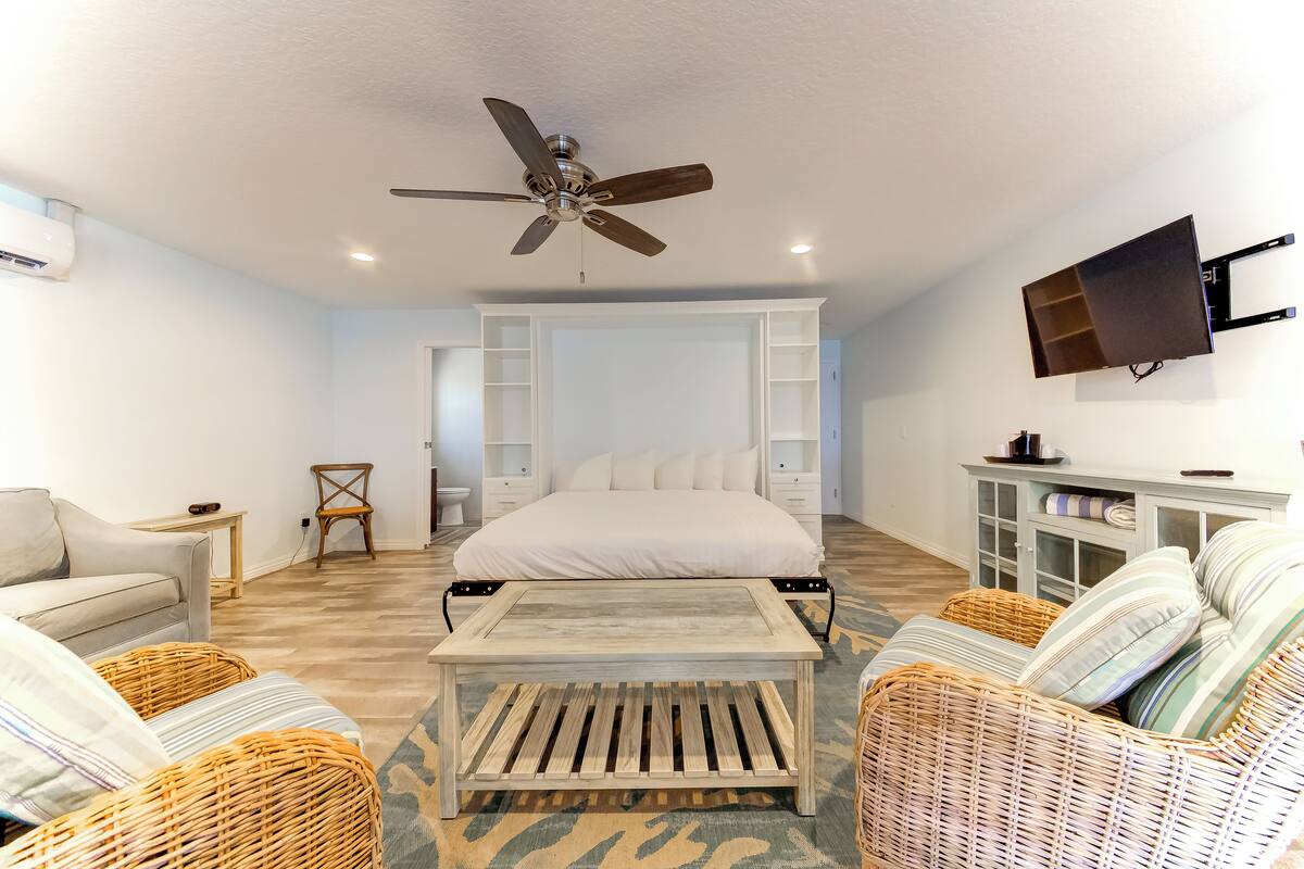 A bedroom with a bed, a ceiling fan, a TV mounted on the wall, wicker furniture, and a small table.