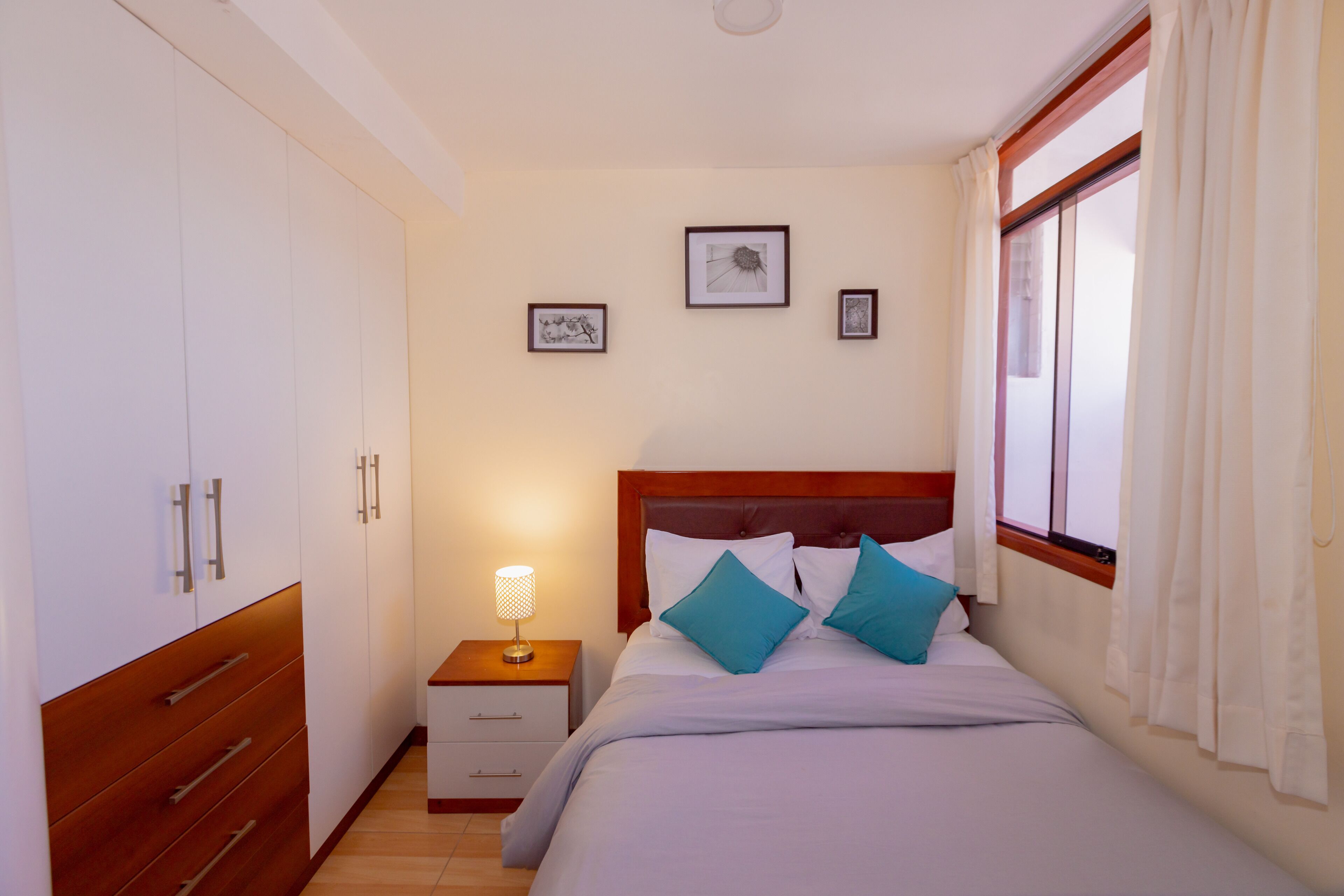 Un dormitorio con cama, mesita de noche, armario y cuadros en la pared.